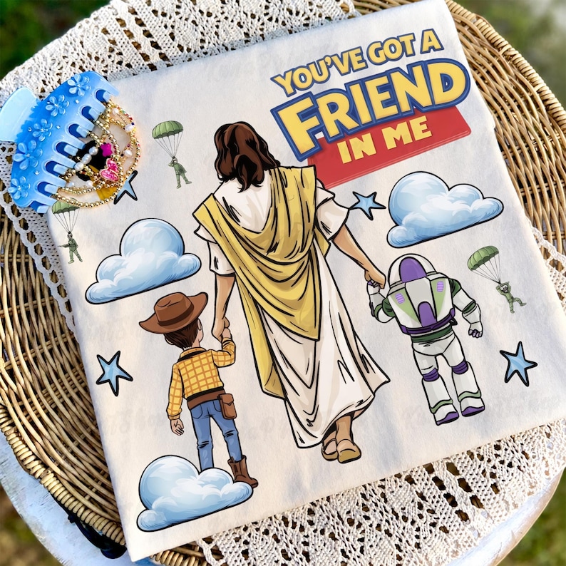 May include: A white t-shirt featuring the text "YOU'VE GOT A FRIEND IN ME" in yellow and red. The shirt has cartoon illustrations of Jesus, Woody, and Buzz Lightyear. Blue clouds, stars, and parachuting figures are also depicted.