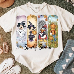 May include: Off-white t-shirt featuring a colorful Halloween design. The design includes cartoon characters dressed in Halloween costumes, such as a ghost, witch, and scarecrow. The words "Spooky Season" are written in a script font.