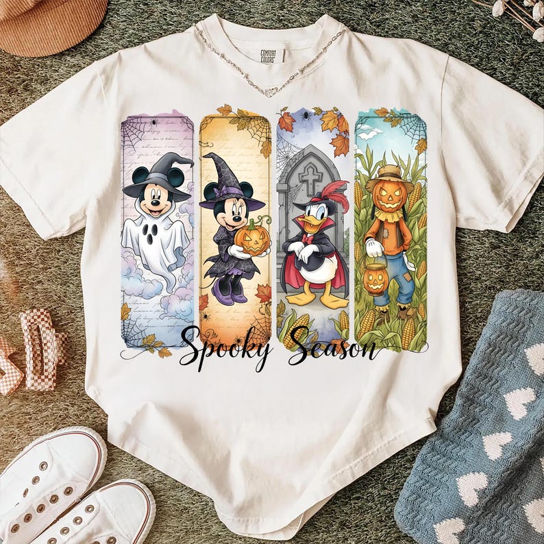 May include: Off-white t-shirt featuring a colorful Halloween design. The design includes cartoon characters dressed in Halloween costumes, such as a ghost, witch, and scarecrow. The words "Spooky Season" are written in a script font.