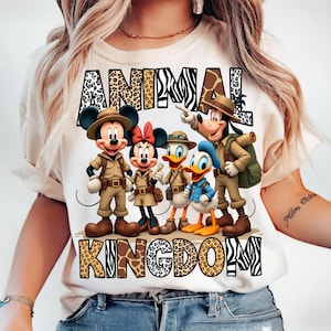 K&ouml;nnte beinhalten: Cremefarbenes T-Shirt mit einer Grafik von Mickey Mouse, Minnie Mouse, Donald Duck, Daisy Duck und Goofy in Safari-Outfits. Die Worte "ANIMAL KINGDOM" sind &uuml;ber und unter den Figuren in Tiermuster gedruckt.