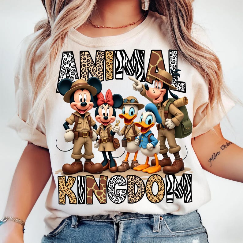 K&ouml;nnte beinhalten: Cremefarbenes T-Shirt mit einer Grafik von Mickey Mouse, Minnie Mouse, Donald Duck, Daisy Duck und Goofy in Safari-Outfits. Die Worte "ANIMAL KINGDOM" sind &uuml;ber und unter den Figuren in Tiermuster gedruckt.