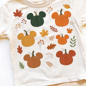 May include: Cream-colored t-shirt featuring a fall-themed design. The design includes pumpkins with mouse ears in various shades of orange, brown, and green, along with autumn leaves and berries. A festive and seasonal graphic.