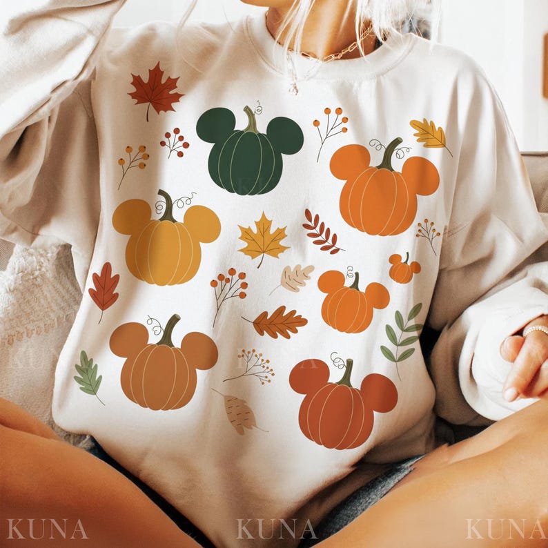 May include: Cream-colored sweatshirt featuring a fall-themed design. The design includes pumpkins with mouse-ear-like shapes in shades of orange, brown, and green, along with autumn leaves and sprigs. The word "KUNA" is printed at the bottom.