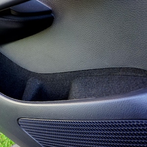 Puede incluir: Primer plano del interior de una puerta de coche, que muestra un compartimento de almacenamiento de tela negra y una rejilla de altavoz negra.