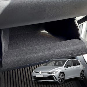 Puede incluir: Un Volkswagen Golf hatchback plateado con molduras negras y llantas negras. El coche está aparcado en un garaje con una alfombra de goma negra en el suelo. El interior del coche es visible, mostrando un compartimento de almacenamiento forrado de tela negra.