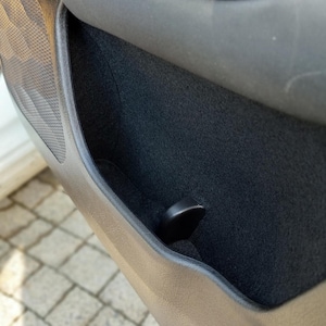 May include: A black car door panel with a storage compartment and a black fabric lining. The compartment has a small black plastic handle.