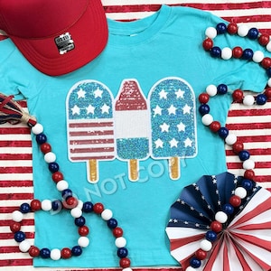 Pode incluir: Uma camiseta azul turquesa com um design de três picolés com lantejoulas. Cada picolé é decorado com estrelas e listras vermelhas, brancas e azuis, que se assemelham à bandeira americana.