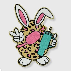 May include: A white bunny with pink ears and a leopard print body is holding a turquoise tumbler with a straw. The bunny is wearing a pink belt with a gold buckle. The bunny is holding a peace sign with its left hand.