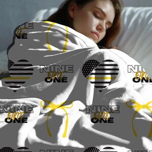 May include: White blanket with a black and white heart design with a yellow stripe. The heart is surrounded by the text "NINE ONE ONE" in black. The blanket also features yellow bows and the text "NINE ONE ONE" repeated throughout.