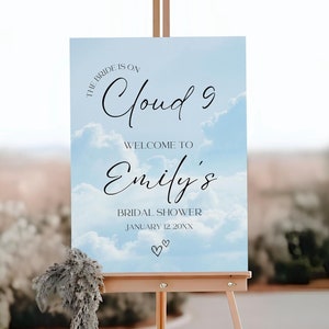 May include: A wooden easel displays a sign with a sky-blue background and white clouds. The sign reads "The Bride is on Cloud 9" and welcomes guests to Emily's Bridal Shower on January 12, 20XX. Two small hearts are at the bottom.