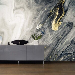May include: A gray and white abstract marble wallpaper with gold accents. The wallpaper is behind a gray cabinet with a black speaker on top.