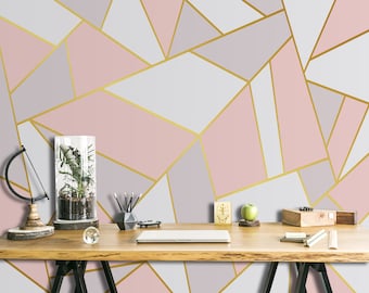 May include: A geometric wallpaper design featuring a pattern of pink, gray, and white shapes outlined with gold lines. A wooden desk with various decorative items, including a terrarium, pencils, and a laptop, is in front of the wallpaper.
