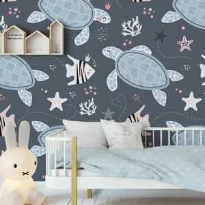 May include: A blue and white nursery wallpaper with a sea life theme. The wallpaper features sea turtles, starfish, coral, and fish. The wallpaper is on a wall behind a white crib with a blue and white blanket.
