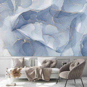 May include: A living room interior with a white couch and a gray armchair. The wall behind the couch is decorated with a blue and gold abstract art print. The couch has a brown throw blanket and two gray pillows. The armchair has a gray pillow.