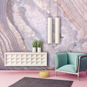May include: A living room interior with a pink wall featuring a large agate-style wallpaper mural. A teal armchair sits in front of the wall, with a white shelf and two potted plants above it. A round yellow ottoman sits on the floor in front of the chair.