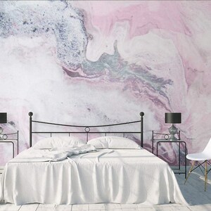 May include: A white bed with a black metal frame sits in front of a pink and gray marbled wall. Two black lamps with white shades are on end tables on either side of the bed. A white chair is in the corner of the room.