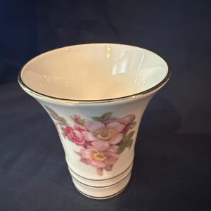 May include: A white ceramic vase with a flared top and a gold rim. The vase features a floral design with pink and green flowers. The base has two black stripes.
