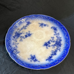 May include: A round, antique porcelain plate with a scalloped edge. The plate features a white center with blue floral designs and a vibrant blue border. Gold accents adorn the rim, adding a touch of elegance to this vintage piece.