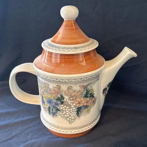 May include: A white ceramic teapot with a brown lid and a floral design featuring grapes, flowers, and leaves. The teapot has a handle and a spout.