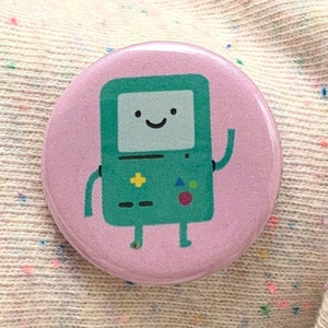 May include: A round, pink button featuring a cartoon character with a green body and a smiling face. The character has a rectangular body with a screen, buttons, and legs. The button is resting on a light-colored sweater.