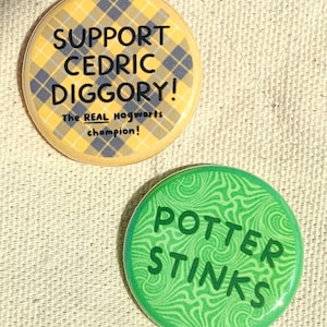 May include: Two round buttons with text and patterns. The first button is yellow with a black and white plaid pattern and the text "SUPPORT CEDRIC DIGGORY! The REAL Hogwarts champion!" The second button is green with a swirling pattern and the text "POTTER STINKS".
