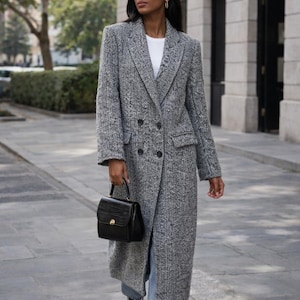 May include: A long, double-breasted gray herringbone coat with a black handbag. The coat has a classic design with large buttons and a tailored fit. The handbag is small and structured. The outfit is completed with light-wash jeans and sneakers.