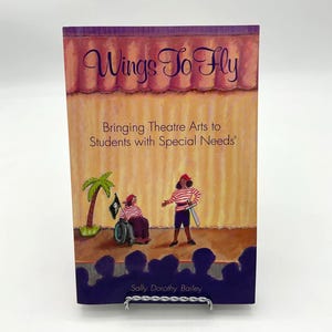 Könnte beinhalten: Ein Buch mit dem Titel "Wings To Fly" und dem Untertitel "Bringing Theatre Arts to Students with Special Needs". Das Cover zeigt eine Bühnenillustration mit zwei Figuren, einer Palme und einer Publikumssilhouette.