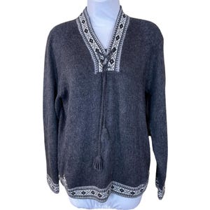 May include: Grey knitted sweater with a white and black geometric pattern around the neckline and hem. The sweater has a tie at the neckline with tassels.