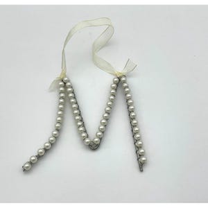 May include: A decorative pearl beaded ornament in the shape of the letter "M". The ornament is made of small, round, white pearls strung on a wire frame. A cream-colored ribbon is attached at the top.