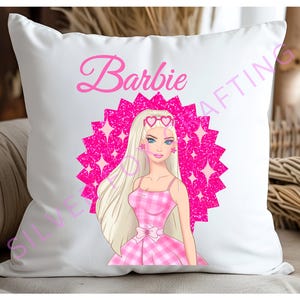 Puede incluir: Almohada blanca con un círculo de purpurina rosa y una imagen de dibujos animados de Barbie con un vestido de cuadros rosa y blanco y gafas de sol. El texto "Barbie" está escrito en rosa encima del círculo.