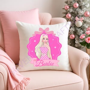 May include: A white decorative pillow featuring a pink graphic of Barbie in a pink polka dot dress. The design includes a pink ribbon, a banner with the name "Barbie", and a pink scalloped frame. The pillow is on a pink chair.
