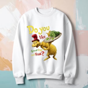 Op de afbeelding: Witte sweatshirt met een cartoon illustratie van een personage dat een dienblad met groene eieren en ham vasthoudt. De tekst "Do you like Green Eggs and Ham?" is gedrukt in geel en groen. Het personage is geel met een rode hoed.