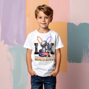 Pode incluir: Uma camiseta branca com um gr&aacute;fico de um trof&eacute;u prateado, o texto "LX" e as palavras "Benito Bowl" em dourado. O design inclui a bandeira de Porto Rico e detalhes coloridos. A camiseta &eacute; usada por um menino.