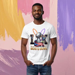 Pode incluir: Camiseta branca com um gr&aacute;fico de um trof&eacute;u prateado, as letras "L X" e as palavras "BENITO BOWL". O design inclui a bandeira de Porto Rico e an&eacute;is coloridos. A camiseta &eacute; usada por uma pessoa.