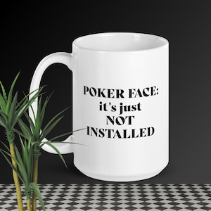 以下が含まれることがあります： 「POKER FACE: it's just NOT INSTALLED」の黒い文字が入った白いセラミックマグ。 マグカップには湾曲したハンドルがあり、白黒のチェック柄の背景に置かれています。 左側には緑の植物が見えます。