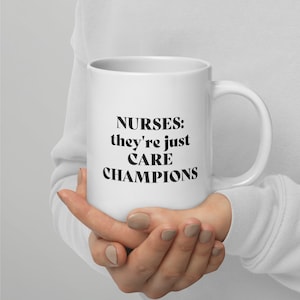 May include: White ceramic mug with the text "NURSES: they're just CARE CHAMPIONS" in black, held by a person. The mug has a curved handle and a glossy finish. The text is in a bold, sans-serif font.