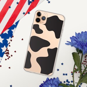 May include: A clear phone case featuring a cow print design with black spots on a beige background. The phone case is surrounded by blue flowers, red heart-shaped confetti, and a patriotic American flag.
