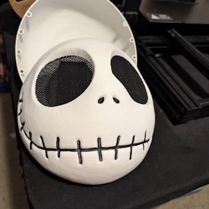 May include: A white, 3D-printed mask resembling Jack Skellington from The Nightmare Before Christmas. The mask features large, black mesh eye openings, a stitched mouth, and a detachable top piece. The mask is designed for cosplay or costume use.