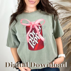 Pu&ograve; includere: Una maglietta verde con un fiocco rosa legato attorno a una lattina di Dr Pepper rossa e bianca.