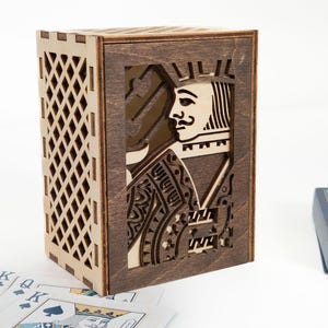 May include: Wooden playing card box with a laser-cut design featuring the King of Spades. The box is rectangular with a diamond lattice pattern on one side and a dark brown finish. Playing cards are visible in the foreground.