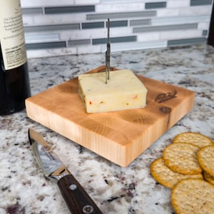 Könnte beinhalten: Ein quadratisches Käsebrett aus Holz mit einem Käseblock und einem Käsemesser. Der Käse hat rote und grüne Flecken. Eine Weinflasche und Cracker sind ebenfalls vorhanden.