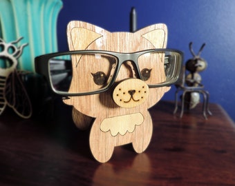 May include: Wooden cat-shaped eyeglasses holder with a pair of glasses resting on it. The cat has a brown and white fur pattern and a cute face with black eyes and a pink nose.