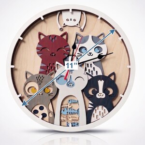May include: A round, white-framed clock with a wooden face featuring a cat theme. The clock is 11 inches in diameter and has five cat figures in various colors. The clock hands are black.