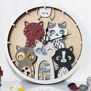 May include: A round wall clock featuring a playful design of six stylized cats in various colors. The clock has a white frame and a light wood-tone background. The cats are in shades of red, gray, black, and white, with blue accents. The clock hands are black and red.