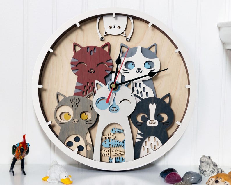 May include: A round wall clock featuring a playful design of six stylized cats in various colors. The clock has a white frame and a light wood-tone background. The cats are in shades of red, gray, black, and white, with blue accents. The clock hands are black and red.