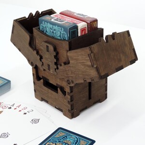 May include: A wooden box with a hinged lid, designed to hold a deck of playing cards. The box is made of dark brown wood and has a simple, elegant design. The lid is open, revealing a deck of cards inside.