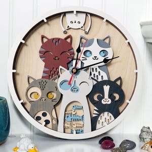 May include: A round wooden wall clock featuring a playful design of several cartoon cats in various colors. The clock has a white frame and a light wood-grain background. The clock hands are black and red.