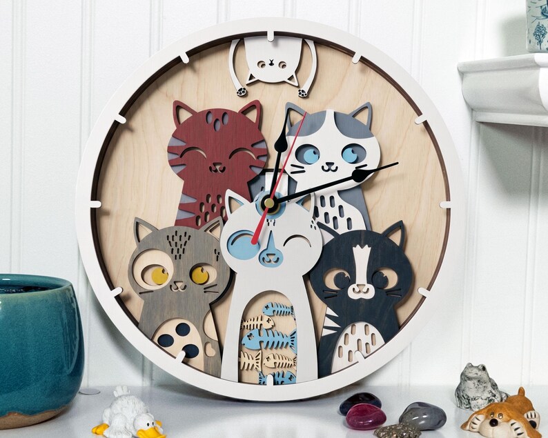 May include: A round wooden wall clock featuring a playful design of several cartoon cats in various colors. The clock has a white frame and a light wood-grain background. The clock hands are black and red.