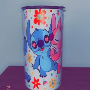 May include: A white tumbler featuring the characters Stitch and Angel from Lilo & Stitch, surrounded by colorful flowers. Stitch is blue, Angel is pink, and the flowers are red, yellow, and orange. The tumbler has a black lid.