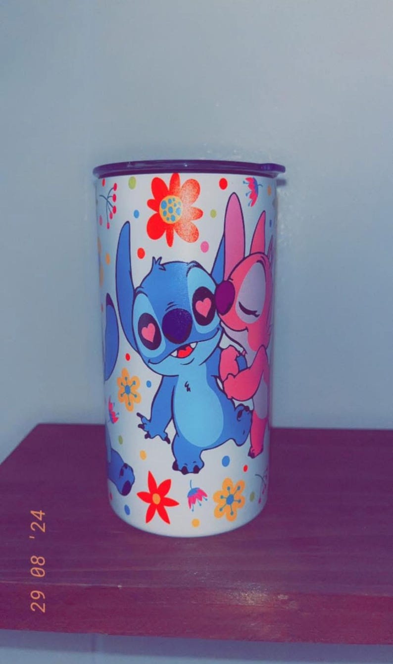 May include: A white tumbler featuring the characters Stitch and Angel from Lilo & Stitch, surrounded by colorful flowers. Stitch is blue, Angel is pink, and the flowers are red, yellow, and orange. The tumbler has a black lid.